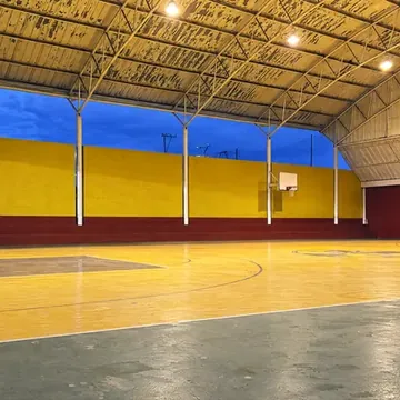 La cancha techada completa al atardecer: piso amarillo con sus líneas, muro amarillo y rojo al fondo, techo en arco y tableros a los lados.